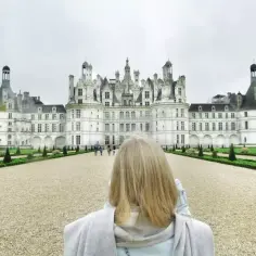 Kobieta z blond włosami podziwia majestatyczne zamki nad Loarą. W oddali widać imponującą architekturę i zadbane ogrody.
