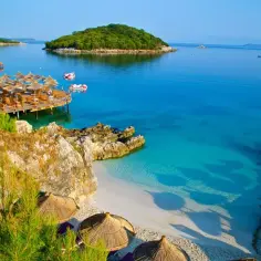 Słoneczny dzień na Ksamil Beach. Drewniany pomost z parasolami, turkusowa woda i zielona wyspa w oddali. Idealne miejsce na relaks.