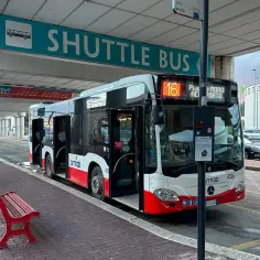 Autobus wahadłowy na lotnisko, z numerem 16, czeka na pasażerów. Czerwona ławka i znak "SHUTTLE BUS" wskazują na punkt przesiadkowy.