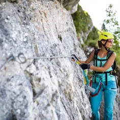 Kobieta w kasku i okularach wspina się po skale, zabezpieczona liną na via ferrata.