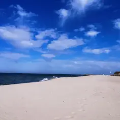 Pusta, piaszczysta plaża nad polskim morzem. Błękitne niebo z białymi chmurami, ciemne morze i wydmy porośnięte trawą.