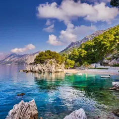 Malownicza riwiera z turkusową wodą, skalistymi zatoczkami i górami w tle. Kilka łodzi kołysze się przy piaszczystej plaży.