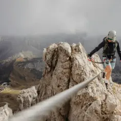 Kobieta w kasku i sprzęcie wspinaczkowym pokonuje via ferrata, balansując na linie nad przepaścią. W tle górzysty krajobraz.