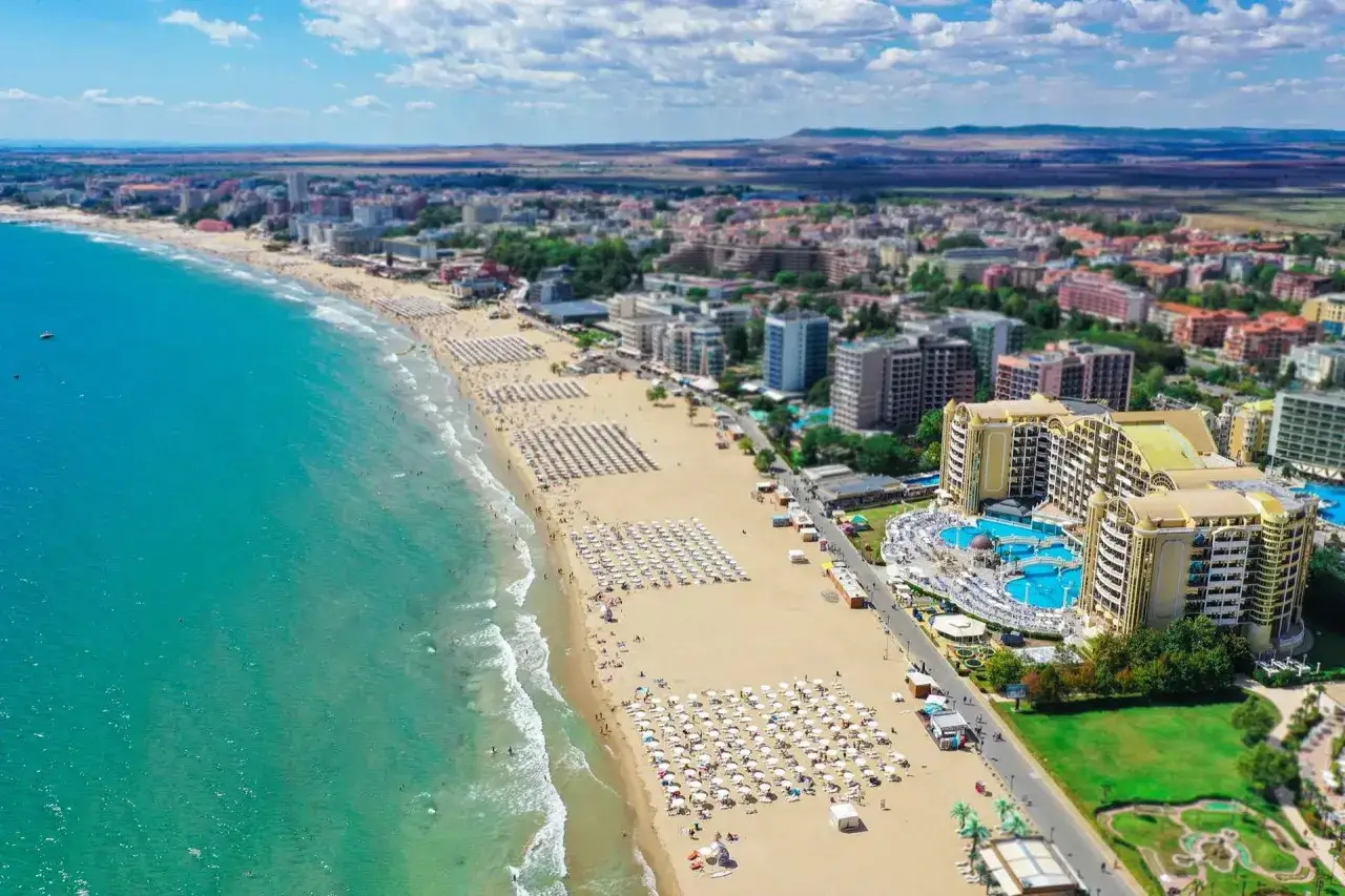 Słoneczna plaża i turkusowe morze – idealne bułgaria wakacje. Tłumy ludzi odpoczywają pod białymi parasolami, a w tle widać hotel z basenami.