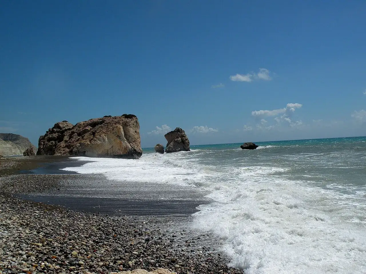 Skała Afrodyty na Cyprze, z falami rozbijającymi się o kamienistą plażę pod błękitnym niebem.