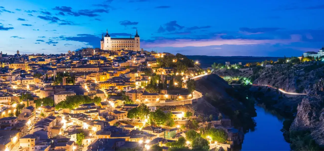 Widok na zabytkowe miasta w Hiszpanii o zmierzchu. Zamek Alcázar w Toledo góruje nad oświetlonymi uliczkami i rzeką Tag.