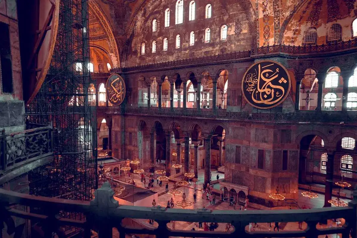 Wnętrze Hagia Sophia z widokiem na kopułę, łuki i ozdobne kaligrafie. Widać ludzi spacerujących po dolnym poziomie.