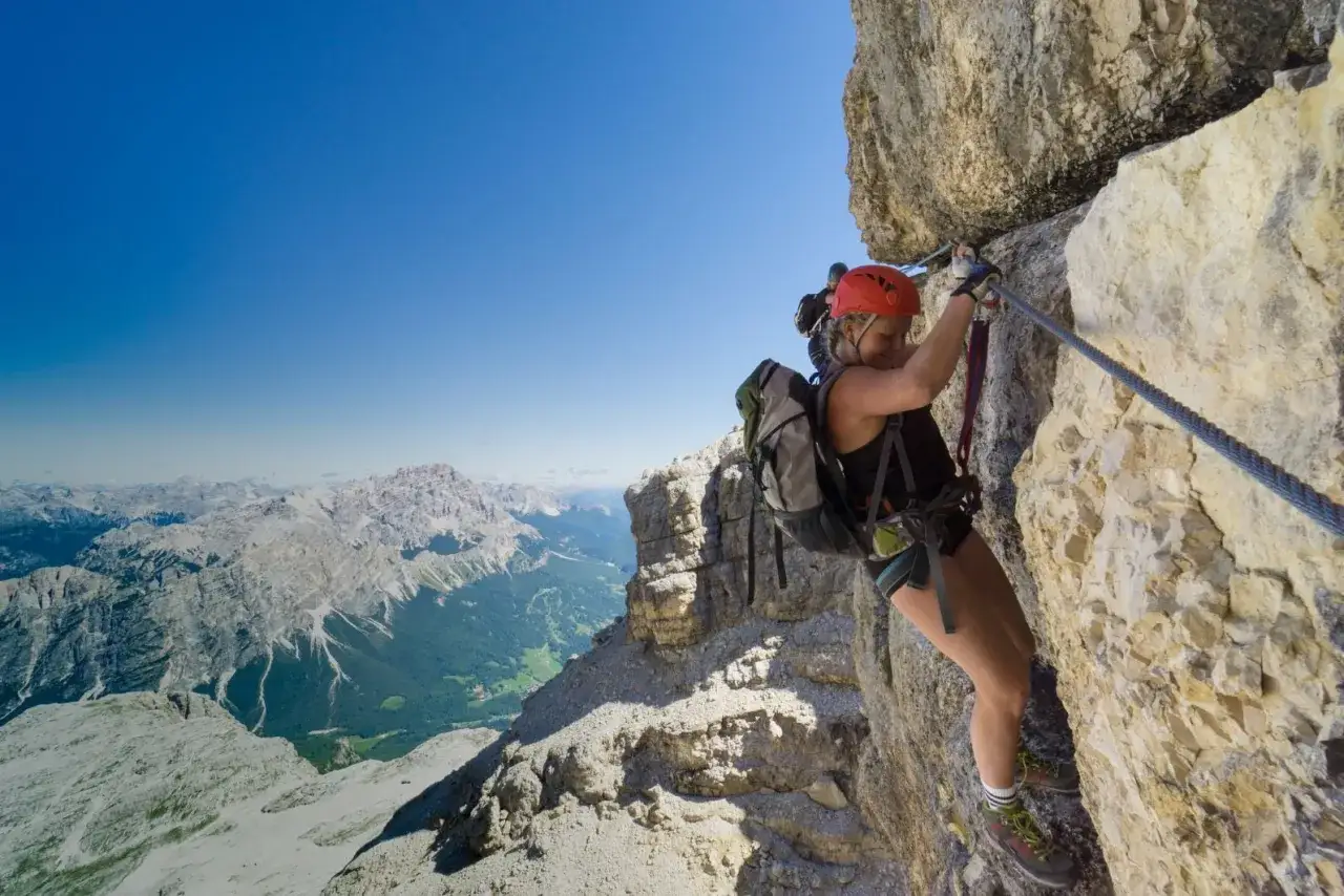 Kobieta w kasku wspina się po skalnej ścianie, korzystając z via ferrata. W tle rozciąga się malowniczy górski krajobraz.
