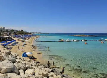 Słoneczna plaża w Protaras z turkusową wodą, łodziami i ludźmi odpoczywającymi na piasku.