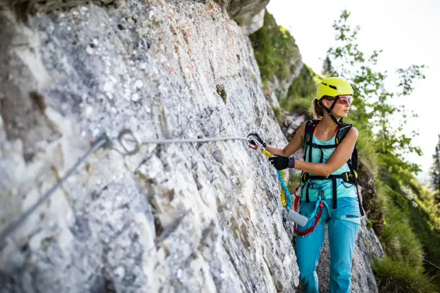 Kobieta w kasku i okularach wspina się po skale, zabezpieczona liną na via ferrata.